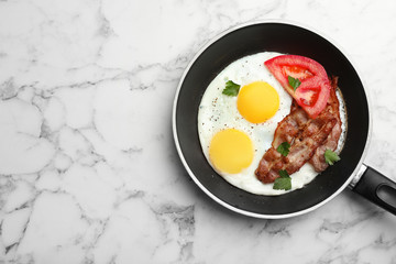 Fried sunny side up eggs with tomato and bacon in pan on marble background, top view. Space for text
