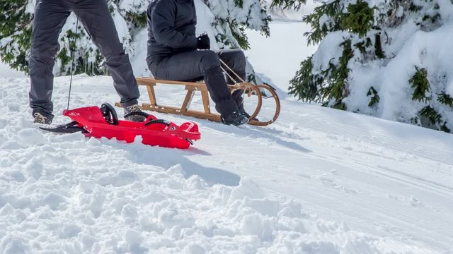 A man is pulling a red sleigh uphill. The other man will sled down the hill before him. It's winter time.