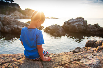 Little boy sits on rocky sea beach at sunset