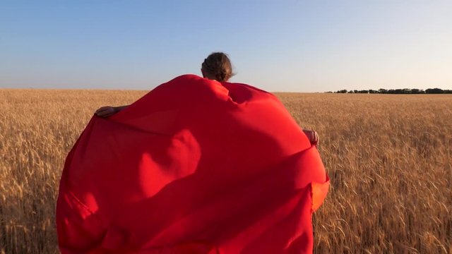 Girl supe rhero running across field with wheat in red cloak against blue sky. Slow motion.