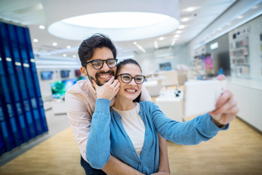 Cute young just married couple in electronic store taking photo with their new phone.Having fun and smiling.