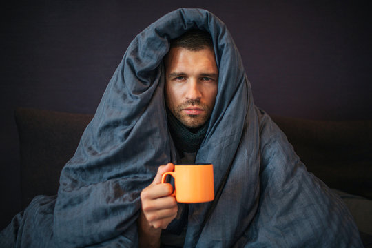 Picture of young man sits on bed and covered up with dark blue blanket. He holds orange cup of tea. Guy looks on camera. He is serious. Young man is emotionless.