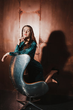 Attractive young woman in a blue green velvet dress, sitting in a leather brown chair, having fun. Long hair, legs, high heels. Luxury concept