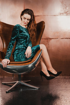 Attractive young woman in a blue green velvet dress, sitting in a leather brown chair, looking down. Long hair, legs, high heels. Luxury concept