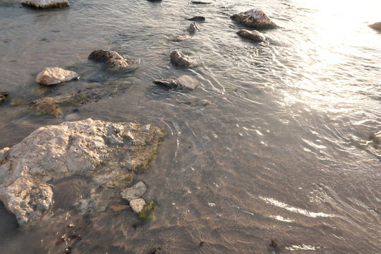 Sea shore with sand and stones and rocks in the water at sunset, close-up