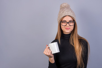 Young beautiful girl with glasses and winter hat standing in front of grey background and holding...