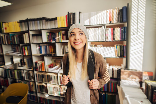 Cute happy school girl standing in library with backpack on her back. Smiling and looking away and satisfied.