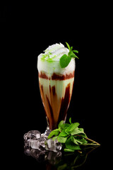 Glass of chocolate milk shake with mint and ice cubes decorated with whipped cream and syrup isolated at black background.