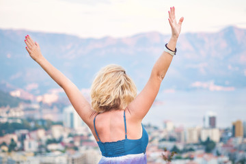 Portrait from the back - a young blonde girl stands on the viewing platform and looks at the city, the seacoast and the mountains, arms outstretched. Concept of freedom and happiness