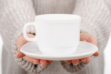 Fototapeta na wymiar Young woman in sweater holding a white cup and saucer in hands, drinking hot coffee on white background. Space for your logo or text.