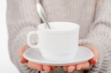 Young woman in sweater holding a white cup and saucer in hands, drinking hot coffee on white background. Space for your logo or text.