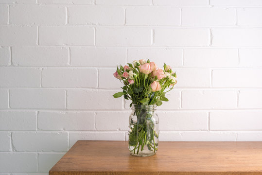 Close up of small pink and cream roses in glass jar on wooden oak sidetable against white painted brick wall