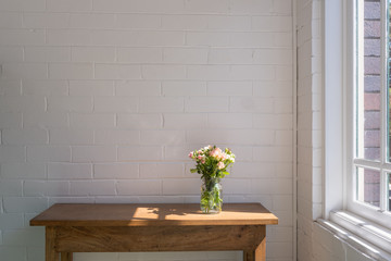 Small pink and cream roses in glass jar on wooden oak sidetable against white painted brick wall...