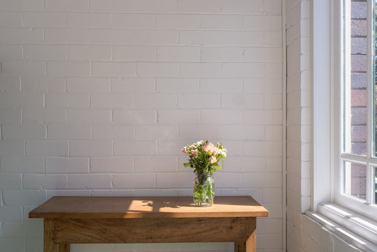 Small pink and cream roses in glass jar on wooden oak sidetable against white painted brick wall next to window with afternoon sunlight