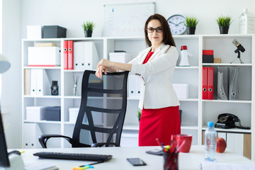 A young girl stands in the office near a chair in a high back.