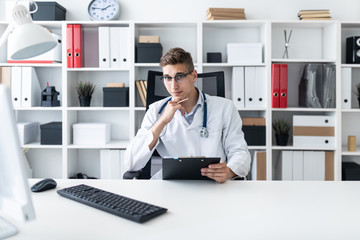 A young man in a white robe sitting at a table in the office. He holds a pen in his hand and looks straight. The portrait of the doctor.