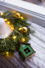 Toy wooden winter house near fir wreath decorated with golden Christmas balls and coiled with glowing garland with warm light on white knitted plaid.