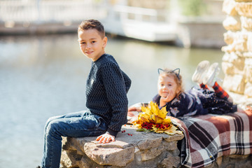 A boy and a girl sitting in a autumn park near a lake on a stone wall with a plaid and yellow leaves
