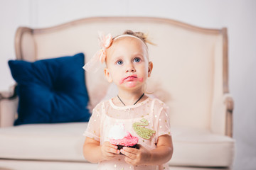 children's birthday. funny two-year-old Caucasian girl in pink dress standing to bedroom of house the background of couch and eating, holding cake dessert. Face and mouth smeared, smeared with cream