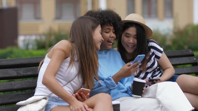 Carefree laughing multi ethnic girls watching funny video content online on cellphone while relaxing on the bench. Joyful female friends resting outdoors with takeaway coffee and sharing smart phone.