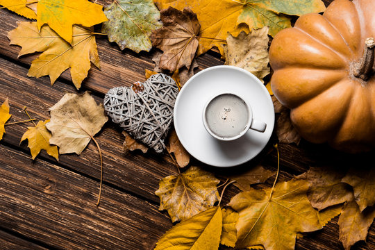 photo of the fallen leaves, pumpkin, heart shaped toy and white cup of coffee on the brown wooden background