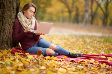 A girl in an autumn park behind a robot on a laptop, on a warm whip and with hot tea. Concept of autumn warmth, atmosphere and comfort, work in nature, freelancing, study