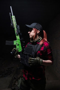 Pretty young woman in body armor with gun/Red-haired girl in a black flak jacket holds a rifle in her hands on a dark background