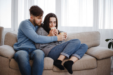 Naklejka na ściany i meble Young couple relaxing at home sitting at sofa with hot drinks. Warming, calmness, home owning, comfort concept