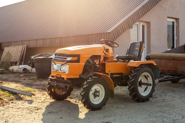 Mini red tractor with trailer standing near hangar building at farm during sunset or sunrise. Small agricultural machinery. Rural country scene