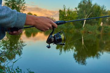 Lake at sunset. A fisherman on the  bank. Fishing rod wheel closeup. Spinning reel. The concept of outdoor activities. Tackles for pike, perch, zander.