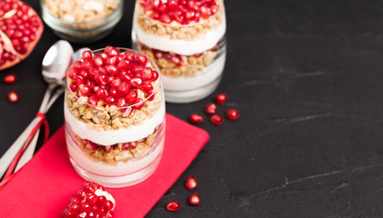 Pomegranate parfait - sweet organic layered dessert with granola flakes, yogurt and fruit seeds on black background.