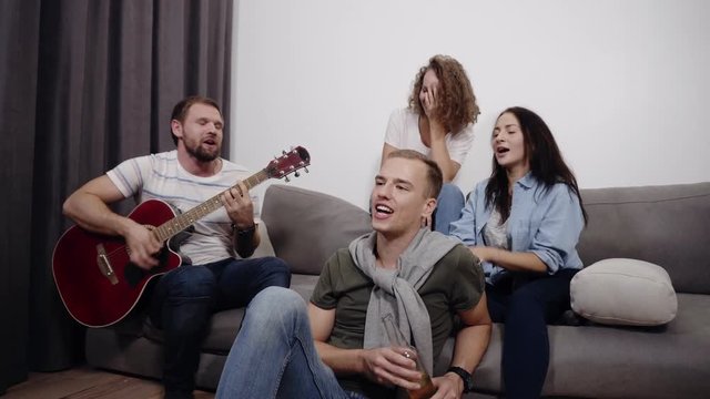 Company of 4 friends sit on sofa and listen to guy playing the acoustic guitar. Get together to have a good time with best friends. Dancing while sitting, singing together, having fun with each other