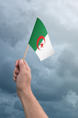 Hand holding Algeria flag high in the air, with a stormy, cloudy sky