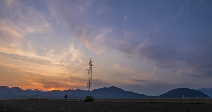 Evening time lapse of a colored orange sunset on an agriculture farming field, that was just tilled, with a power electricity line in the middle and hills in the background.