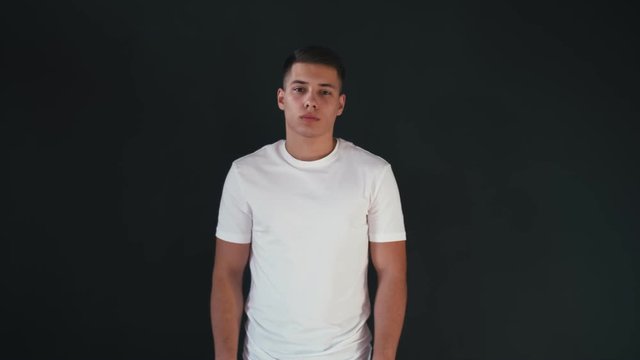 Handsome and positive young man stands in white shirts and looks on camera. Guy moves head from one side to another. He starts to smile and change emotion to playful. Isolated on black background