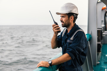 Marine Deck Officer or Chief mate on deck of vessel or ship . He holds VHF walkie-talkie radio in hands. Ship communication - obrazy, fototapety, plakaty