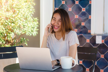 Cheerful asian young woman sitting in cafe drinking coffee and using smartphone for talking, reading and texting. Attractive asian woman holding a cup of coffee while working on laptop.