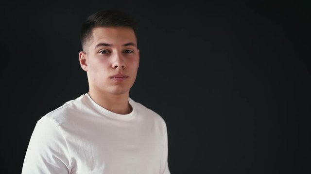 Young man stands on left side and looks on camera. He is serious. Suddenly guy stads to laugh. In the end he makes funny emotion on his face. Isolated on black background.