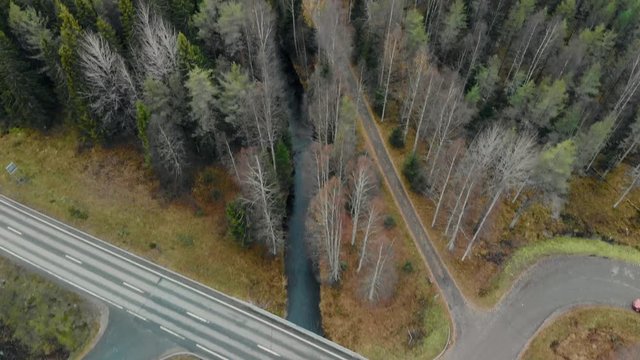 Aerial, tilt, drone shot, above a river and a ashalt road bridge, between pine trees and leafless birch forest, on a cloudy, autumn day, in Juuka, North Karelia, Finland