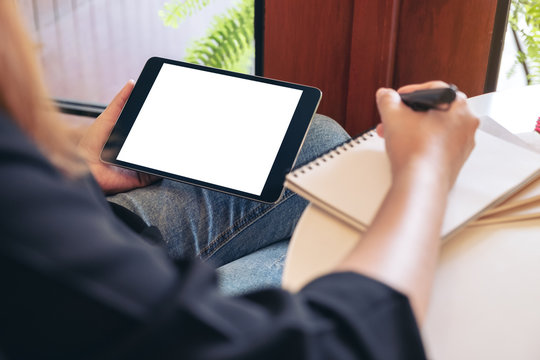 Mockup image of a woman holding and using black tablet pc with blank white desktop screen while writing on notebooks in office