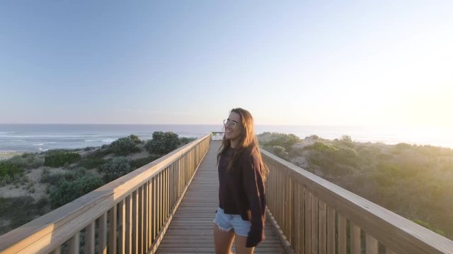 Follow shot of young girl smiling and walking along jetty at beach during sunset,