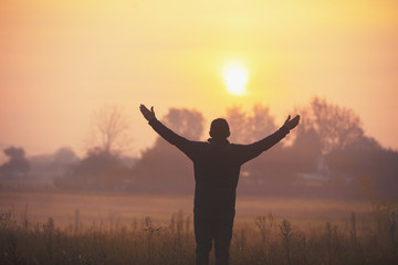 A happy man with hands in the air standing in the field in the early morning and looking at a sunrise