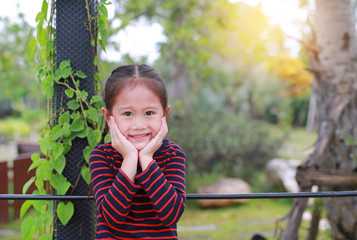 Portrait of smiling little Asian kid girl with touching her cheek looking straight at camera in the nature garden.