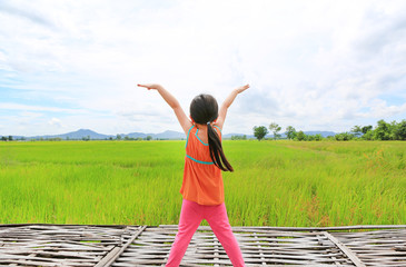 Rear view of little Asian child girl stretch arms and relaxed at the young green paddy fields with mountain and cloud sky.