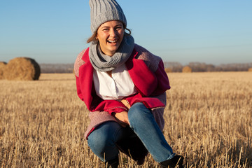 Naklejka na ściany i meble Portrait of a model in gray knitted hat and warm clothes have fun, sitting on field in sunny autumn day . Autumn warm photo. Woman smiling and look at camera, joyful cheerful mood.