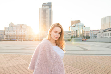 Pretty girl in a pink coat poses on the camera at the background of the street at sunset. Model in...