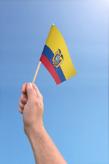 Hand holding Ecuador flag high in the air, with a clear blue sky