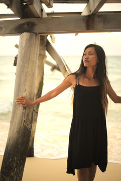 Young asian Vietnamese woman standing beneath old wooden pier on beach posing with sea in background