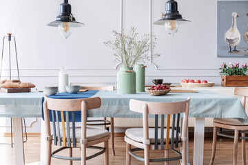 Wooden chairs at table with flowers and food in white cottage dining room interior with lamps and poster. Real photo