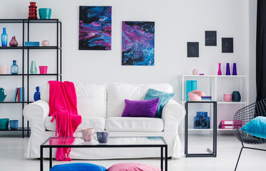 Metal shelves with vases behind white couch with pink blanket and purple and blue pillows, real photo